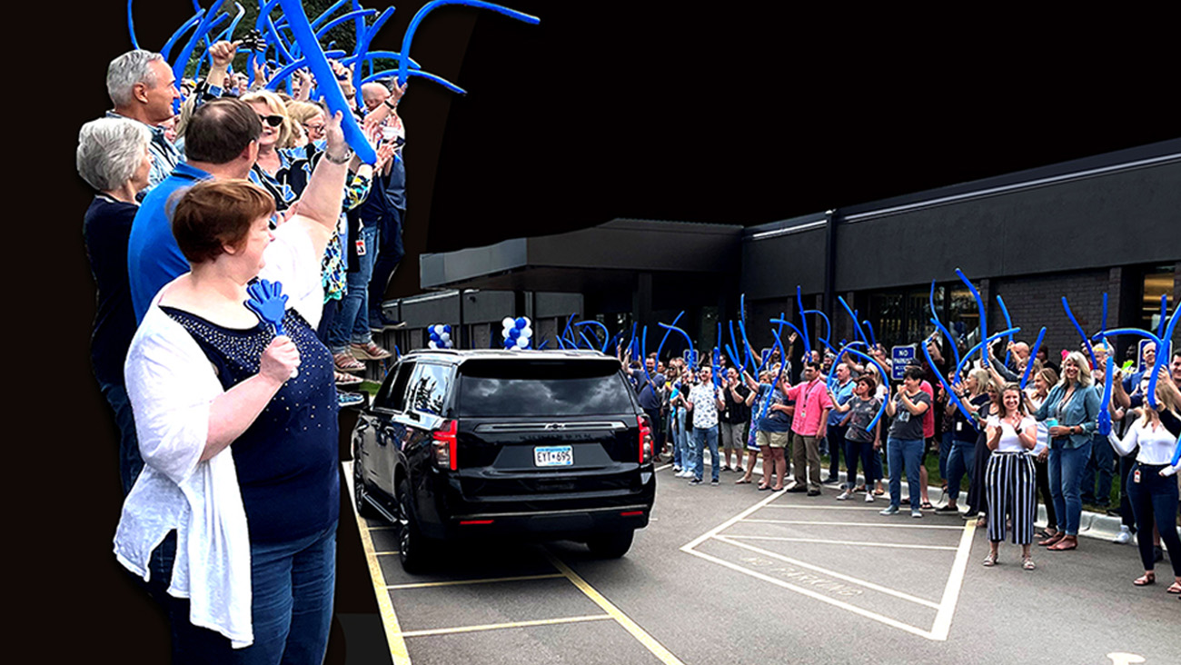 A large crowd stands outside a building, waving blue inflatable sticks as a black SUV drives by. The people appear to be organized on either side of a driveway, cheering and celebrating as the vehicle passes.