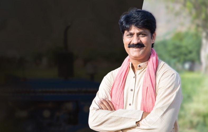 A man with a mustache stands confidently with arms crossed, wearing a light-colored shirt and a pink scarf. The background is blurred, featuring greenery and a dark area to the left.