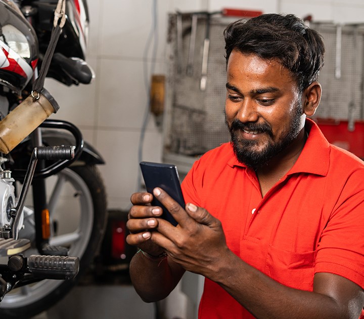 A man in a red shirt is smiling while looking at his phone in a workshop. He stands near a motorbike, with tools hanging on the wall behind him.