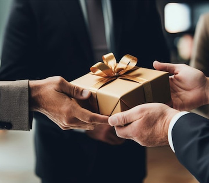 Two people in formal attire exchanging a wrapped gift with a gold ribbon. The focus is on their hands and the gift, creating a sense of anticipation and celebration.