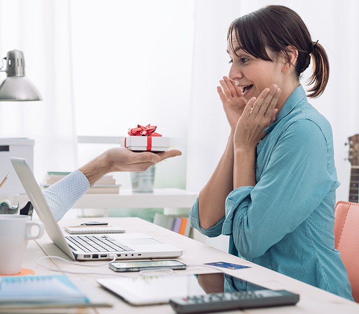 A surprised woman in a blue shirt sits at a desk, covering her mouth with her hands in delight. An arm emerging from a laptop screen presents her with a small gift box adorned with a red ribbon. The scene suggests virtual gift-giving.