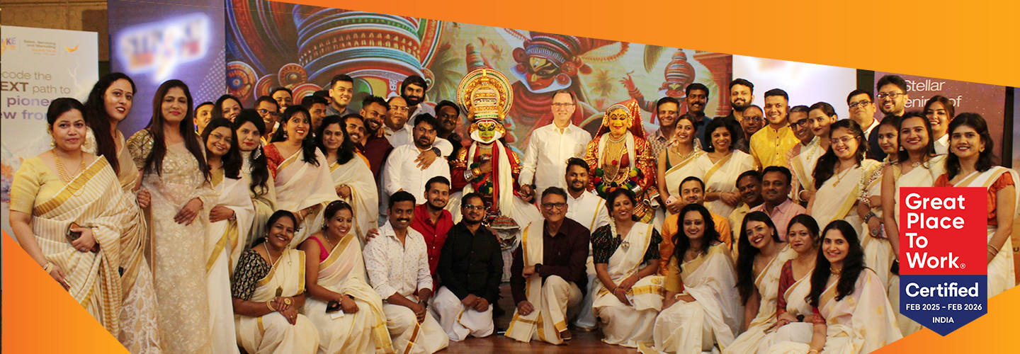 A large group of people dressed in traditional attire pose together, celebrating at an event. Theres a colorful backdrop with Indian motifs and a Great Place to Work certification logo in the corner.