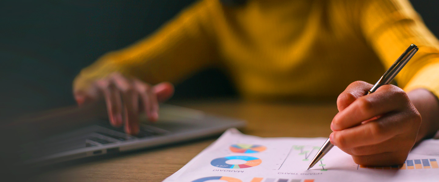 A person in a yellow sweater is writing on a financial report with charts and graphs, using a pen. Their other hand is on a laptop keyboard. The background is blurred.
