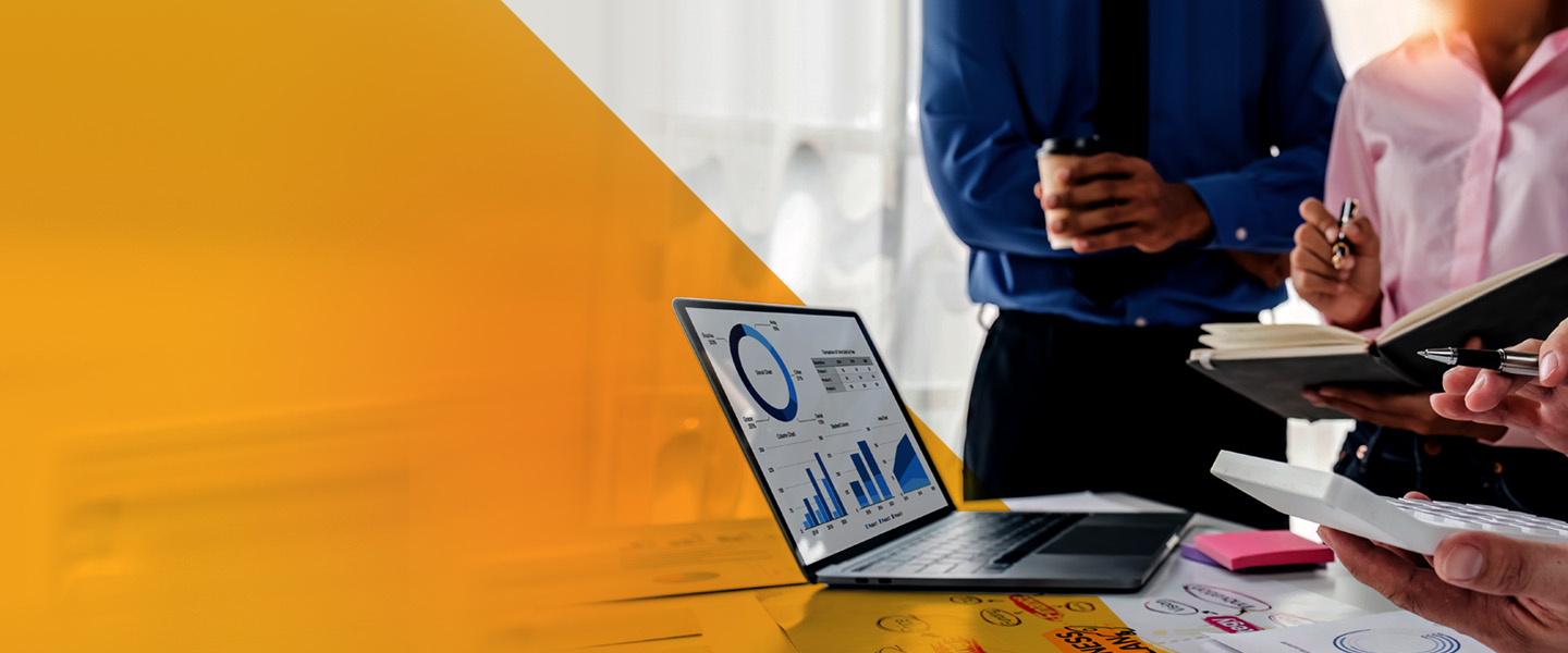 Two people stand at a table with a laptop displaying graphs. Papers with sketches and a calculator are on the table. One person is holding a notebook, and the other a coffee cup. The left side of the image features a yellow geometric shape.