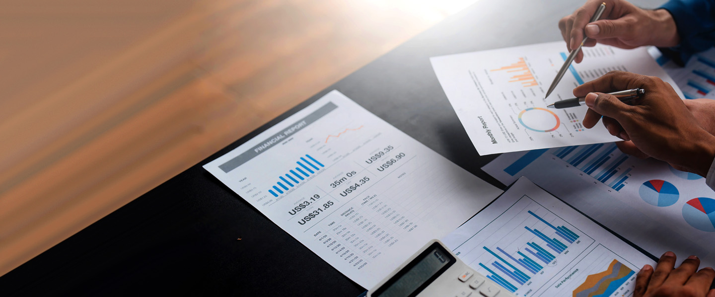 Close-up of hands holding pens over financial documents with charts and graphs. A calculator is on the table. The scene suggests a collaborative business or financial planning meeting.