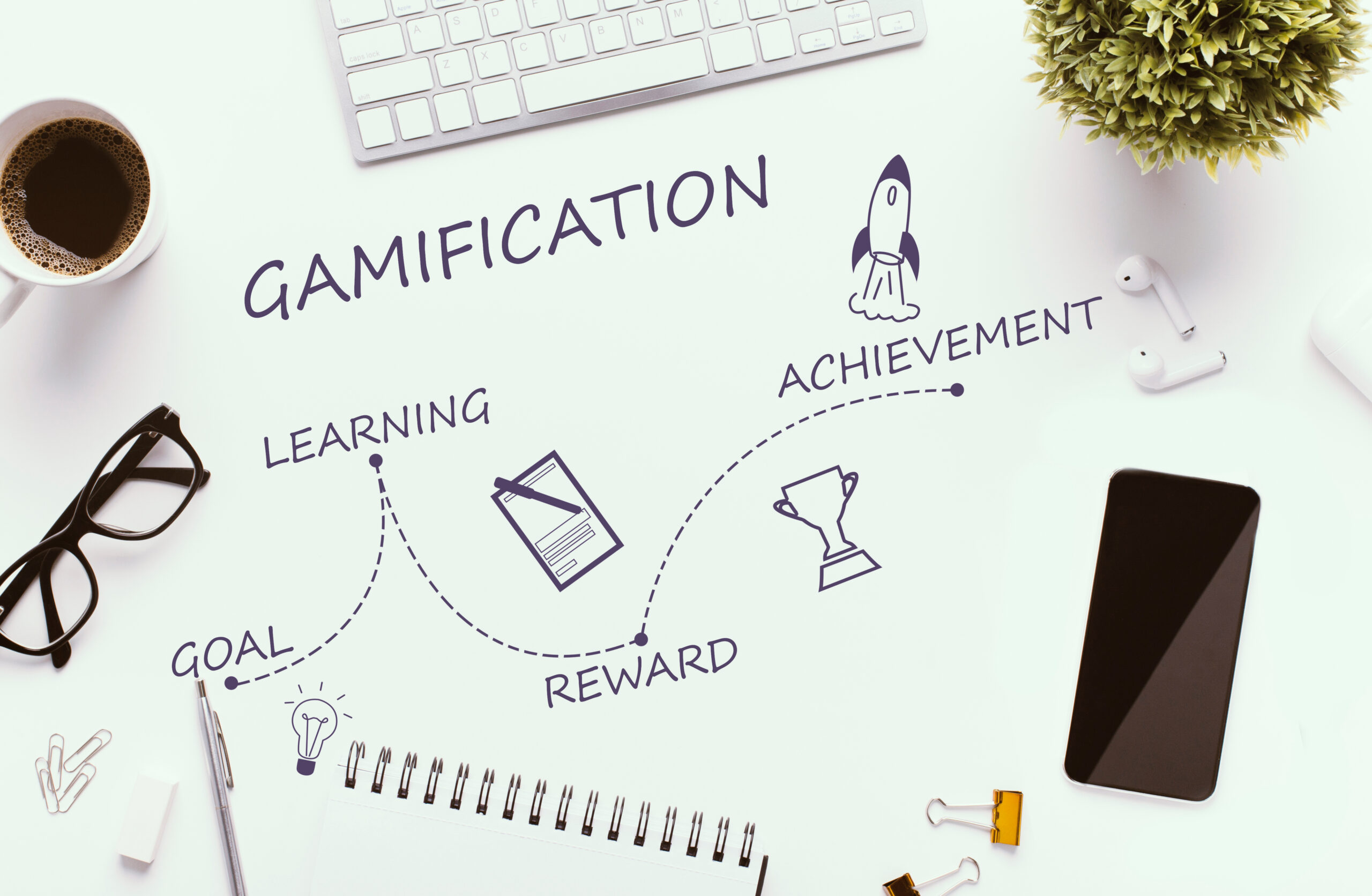 A desk with a keyboard, coffee, plant, glasses, earbuds, and a smartphone. The word Gamification is written above icons and words: Goal (lightbulb), Learning (notebook), Reward (rocket), Achievement (trophy).