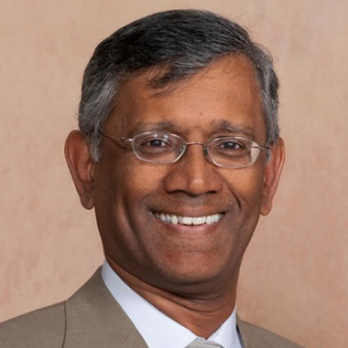 Smiling man with short gray hair and glasses, wearing a brown suit. Neutral, light brown background.