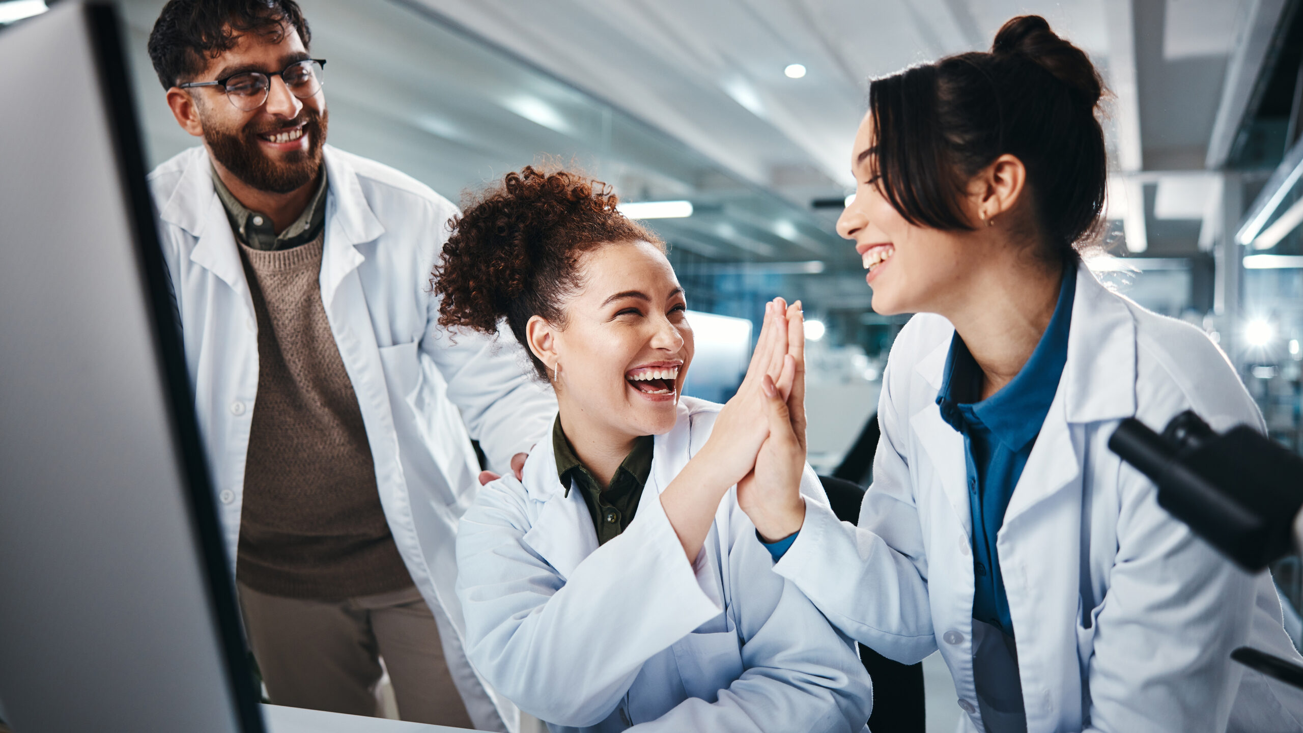 Three scientists wearing lab coats celebrate in a lab. Two women high-five joyfully, while a man with glasses smiles at them. They are gathered around a computer, creating a cheerful and collaborative atmosphere.