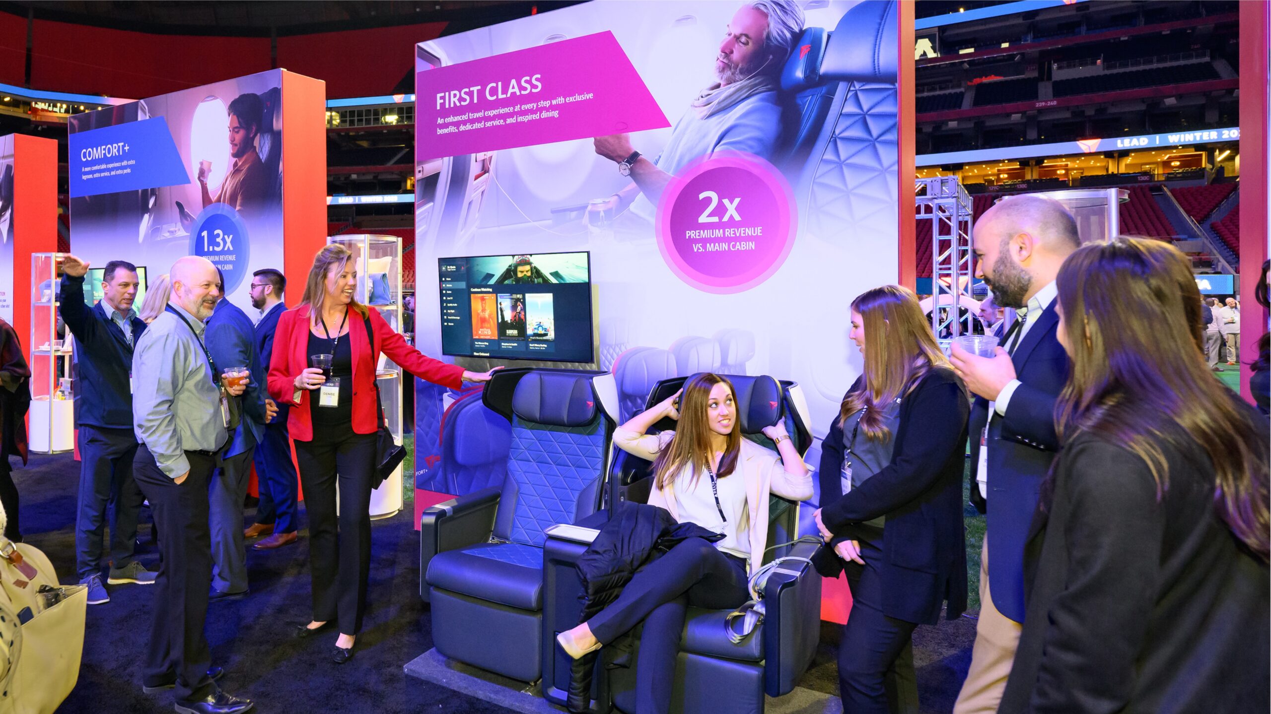 People are gathered around a display booth showcasing first-class airline seats at an exhibition. A woman is sitting in one of the seats, demonstrating its features, while others stand around conversing and observing.