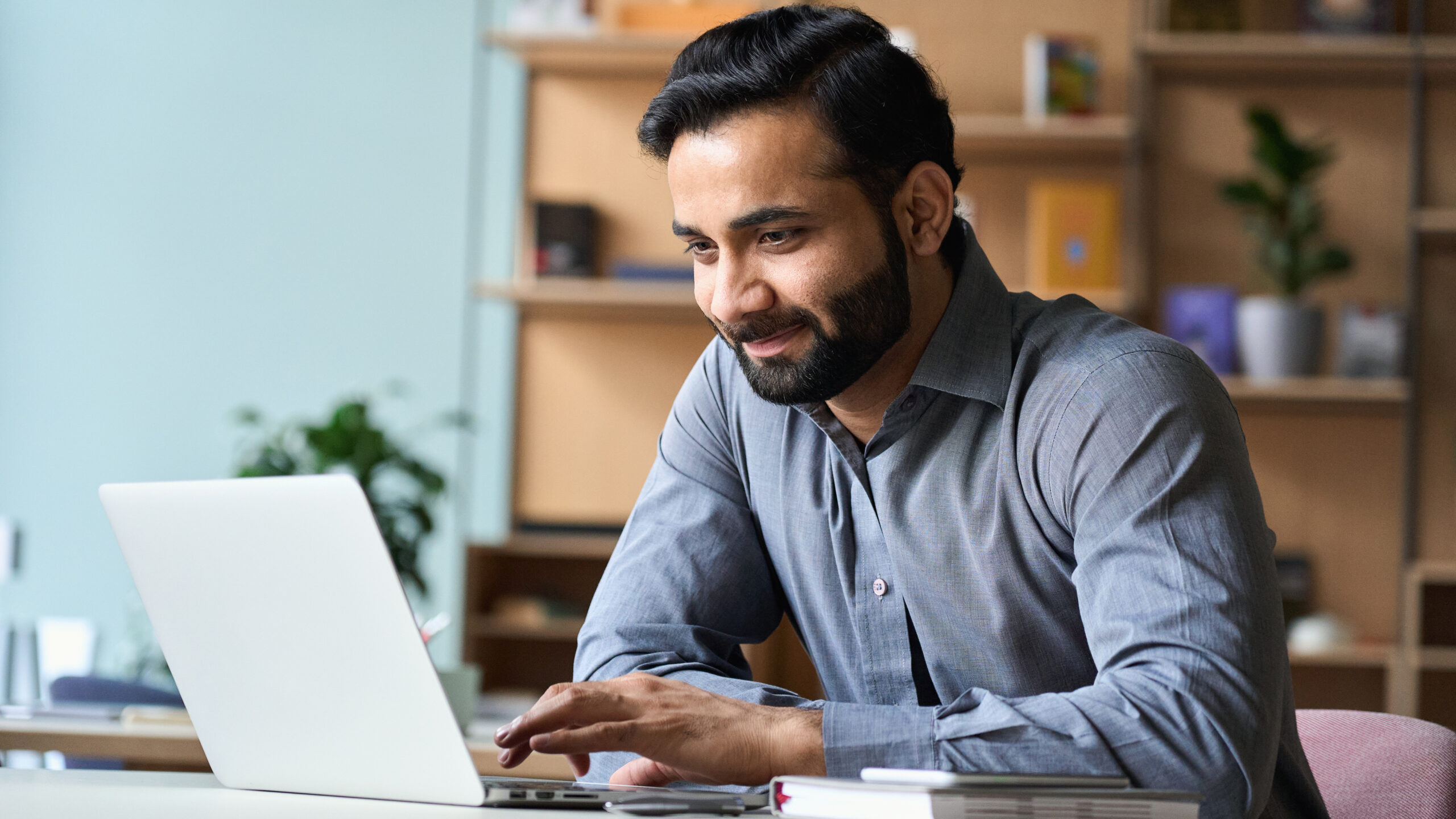 A man in a gray shirt is smiling while working on a laptop at a desk. Behind him are shelves with books and a plant, creating a comfortable workspace atmosphere.