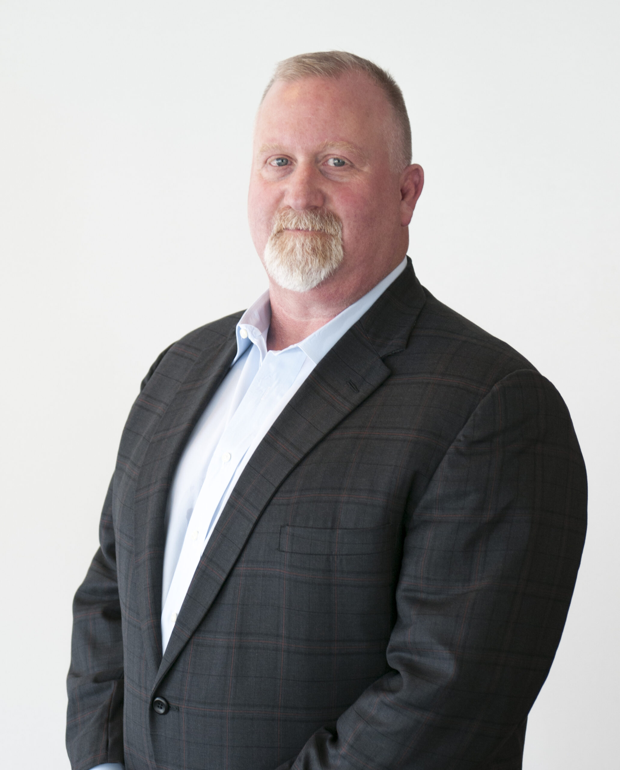 A man with a short beard and mustache, wearing a plaid suit jacket and a light blue shirt, stands against a plain white background, looking at the camera with a neutral expression.
