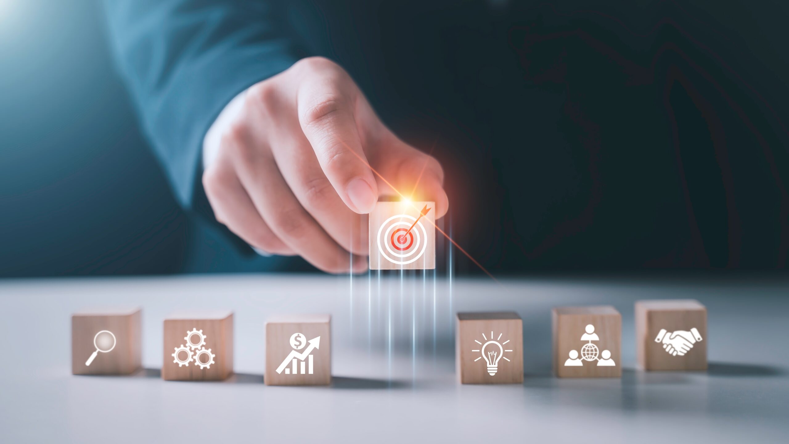 A hand touches a glowing target icon above a wooden cube, flanked by cubes with icons representing search, settings, growth, idea, and handshake, symbolizing strategy and goal setting in business.