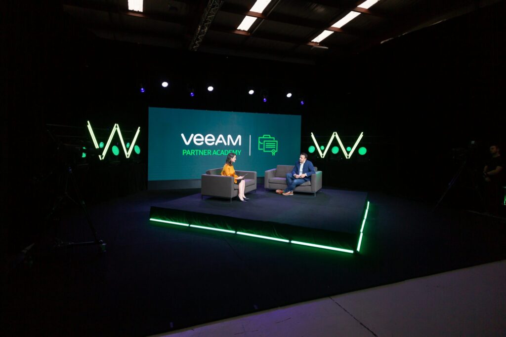 Two people sit on a stage with Veeam Partner Academy displayed on a large screen behind them. The stage is lit in green, and there are two dark chairs. The setting appears to be a professional event or presentation.