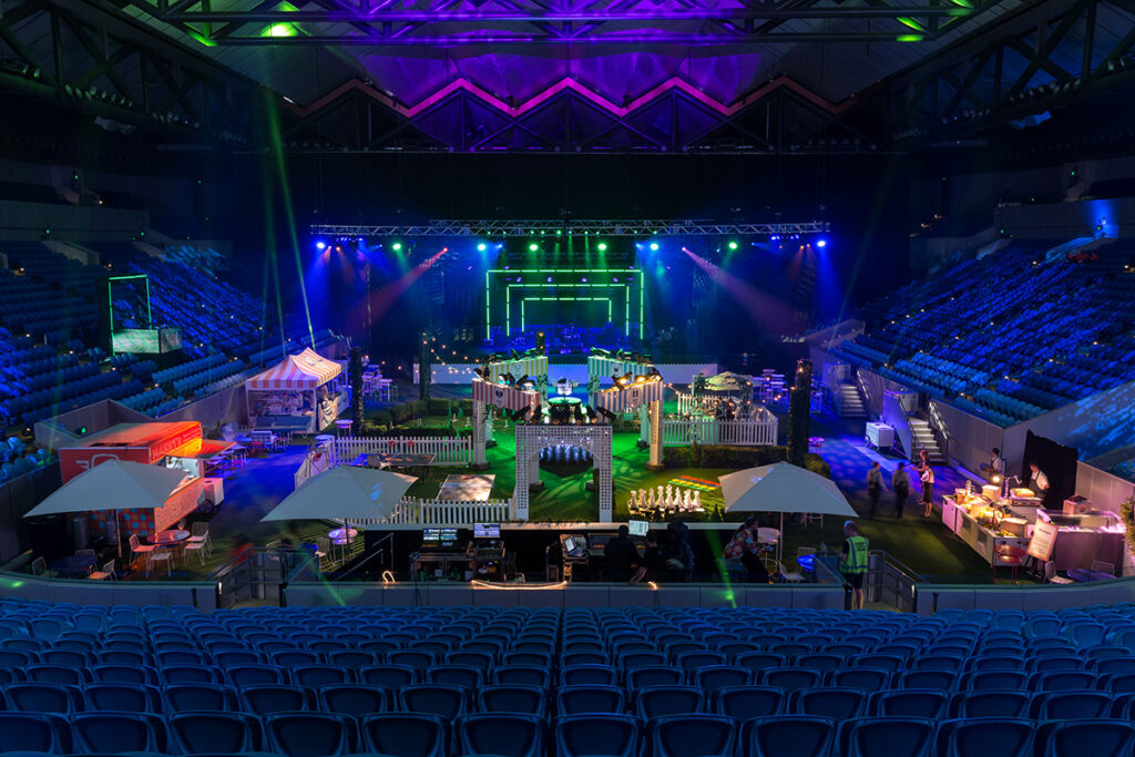 A large indoor arena transformed into a colorful event space. Booths with lights and umbrellas are scattered across the floor. Vibrant purple and green spotlights illuminate the stage, set against rows of empty blue seats.