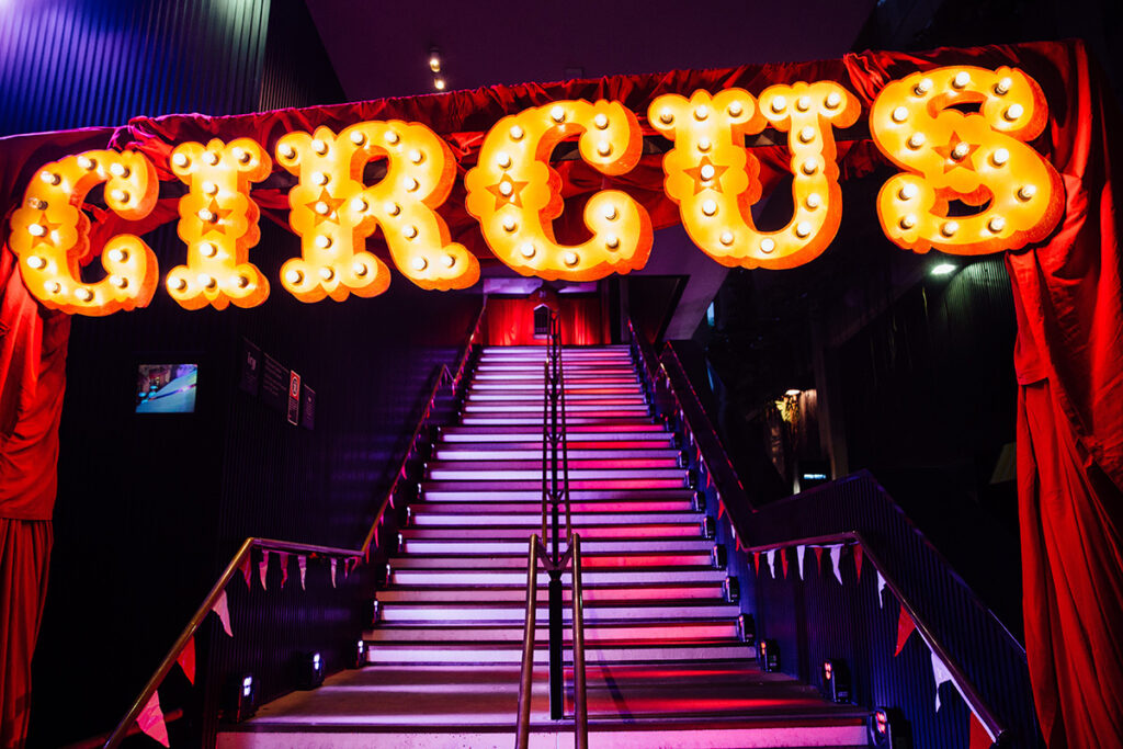 Illuminated Circus sign made of large, vintage-style light bulbs hangs above a stairway decorated with red drapes and triangular flags. The colorful lighting creates a festive and inviting atmosphere.