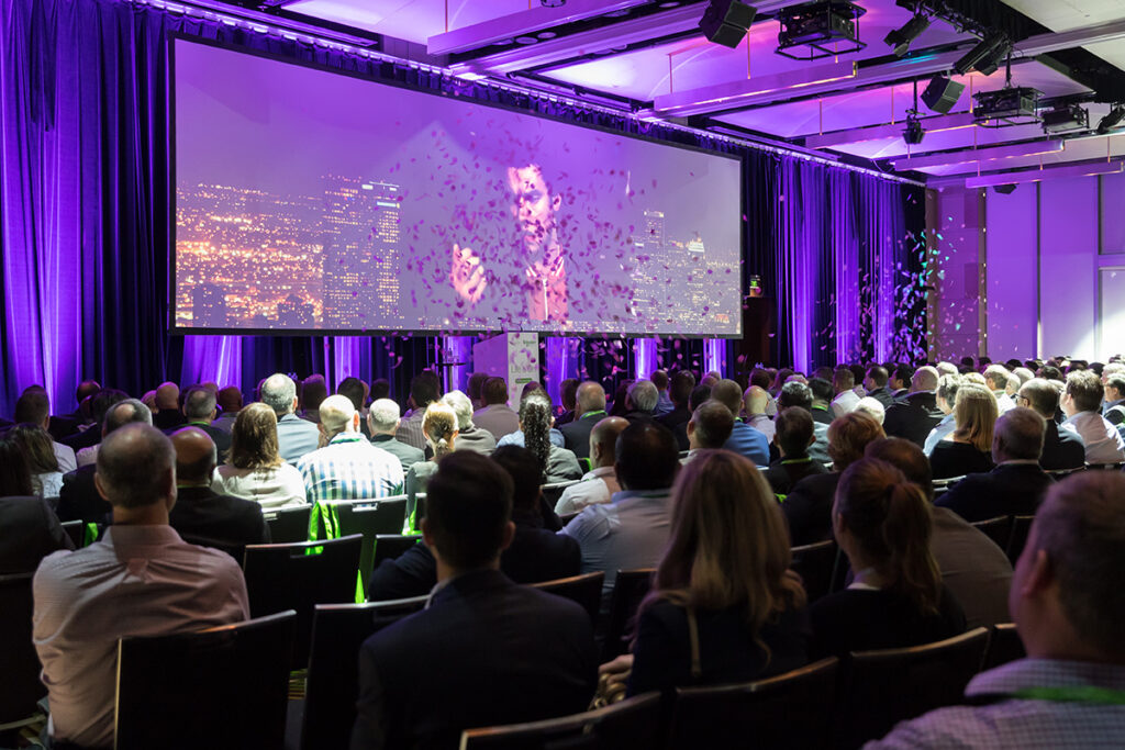 Audience seated in a large conference room, watching a presentation on a screen. The room is illuminated with purple lighting, and confetti is falling from above. The audience is facing away from the camera.