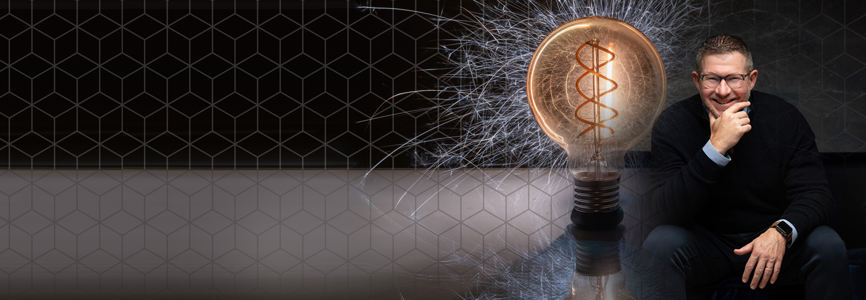 A man in glasses and a dark sweater sits smiling with his chin resting on his hand. Next to him is an illuminated light bulb, with visible filament resembling DNA, surrounded by sparks. The background features a geometric pattern.