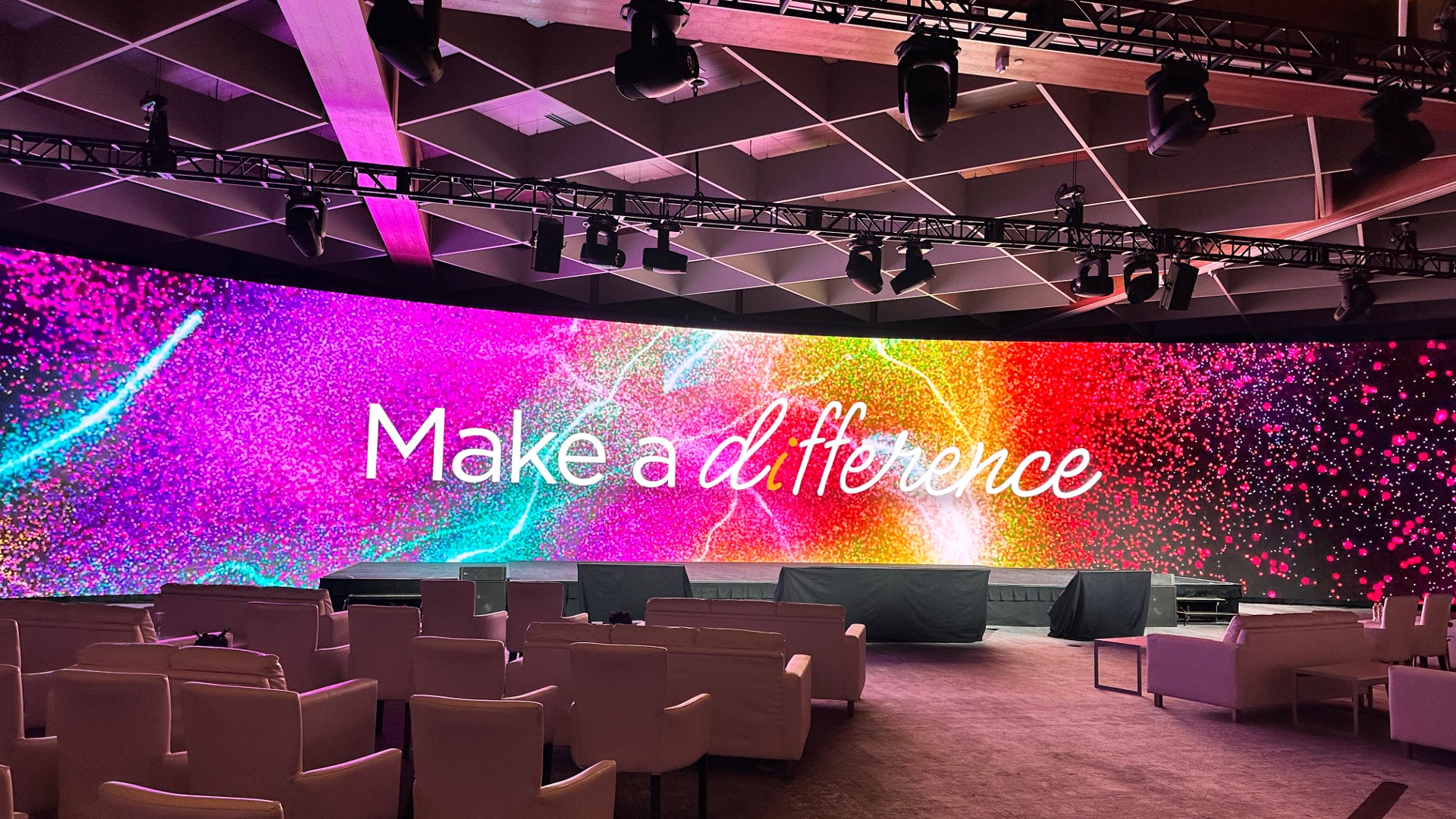 Large LED screen displays Make a Difference in elegant script over a vibrant, multicolor background. White chairs are arranged in rows facing the screen, under a ceiling with spotlights and beams.