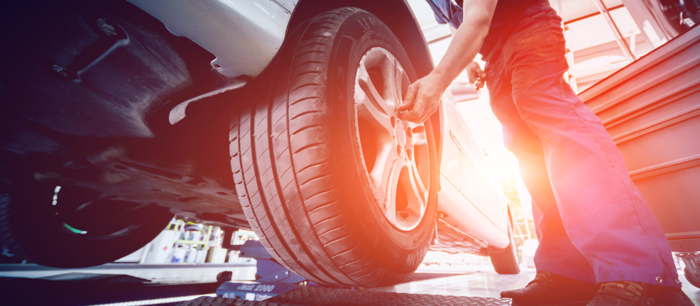 A mechanic in red overalls works on a cars wheel, adjusting the lug nuts in a sunlit garage. The vehicle is elevated on a lift, and the scene is filled with bright light and workshop equipment.