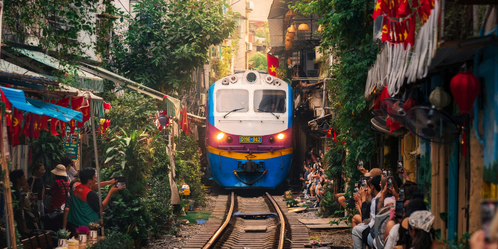 A train travels through a narrow, vibrant street surrounded by buildings in Hanoi. People on either side take photos and observe. The area is adorned with greenery and red flags, creating a lively and bustling atmosphere.