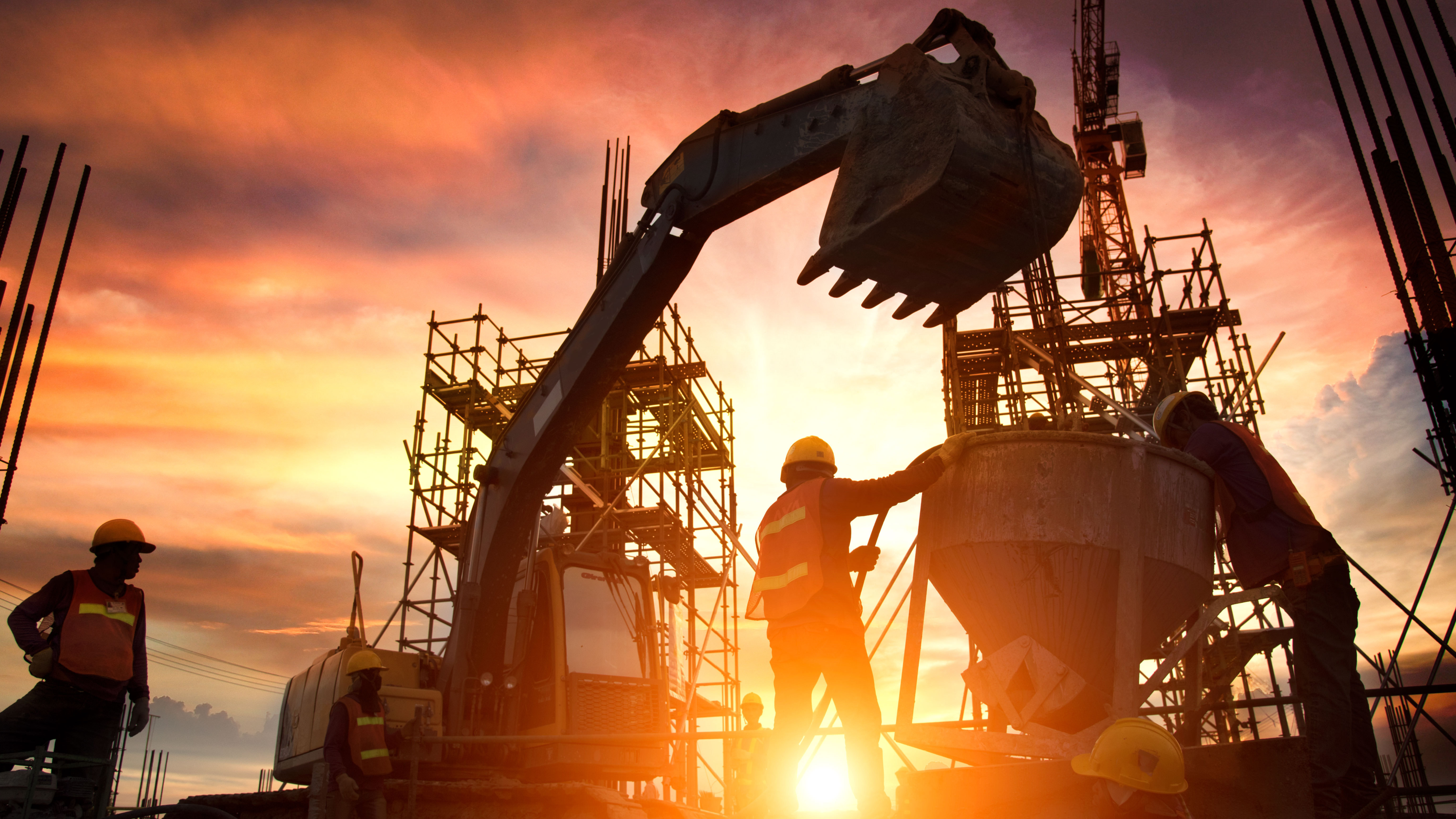 Silhouetted construction workers and machinery on a site with scaffolding, set against a vibrant sunset sky. The sun is low on the horizon, casting a warm glow and creating dramatic lighting effects.