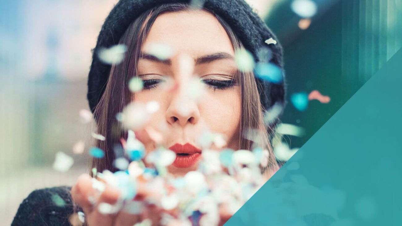 A woman wearing a knit hat blows a handful of colorful confetti towards the camera, creating a lively and festive atmosphere. The background is softly blurred, emphasizing the flying confetti and her joyful expression.