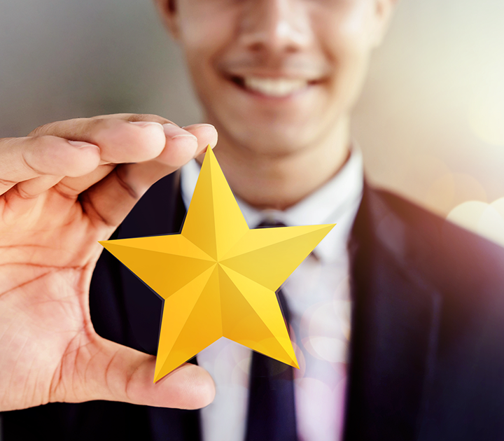 A person in a suit smiling while holding a golden star between their thumb and index finger. The image focuses on the star, with a blurred background giving a sense of brightness and celebration.