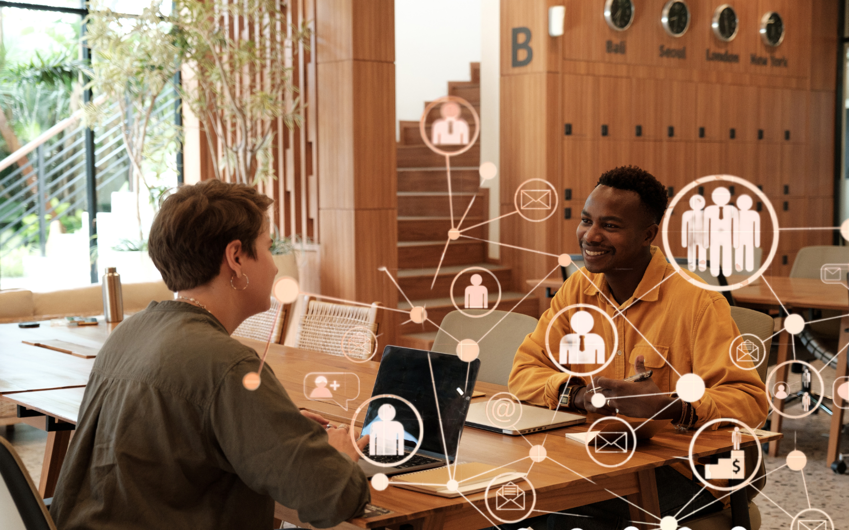 Two people sit across from each other at a wooden table in a modern office, smiling and talking. Digital icons float in the foreground, representing technology and social connections.
