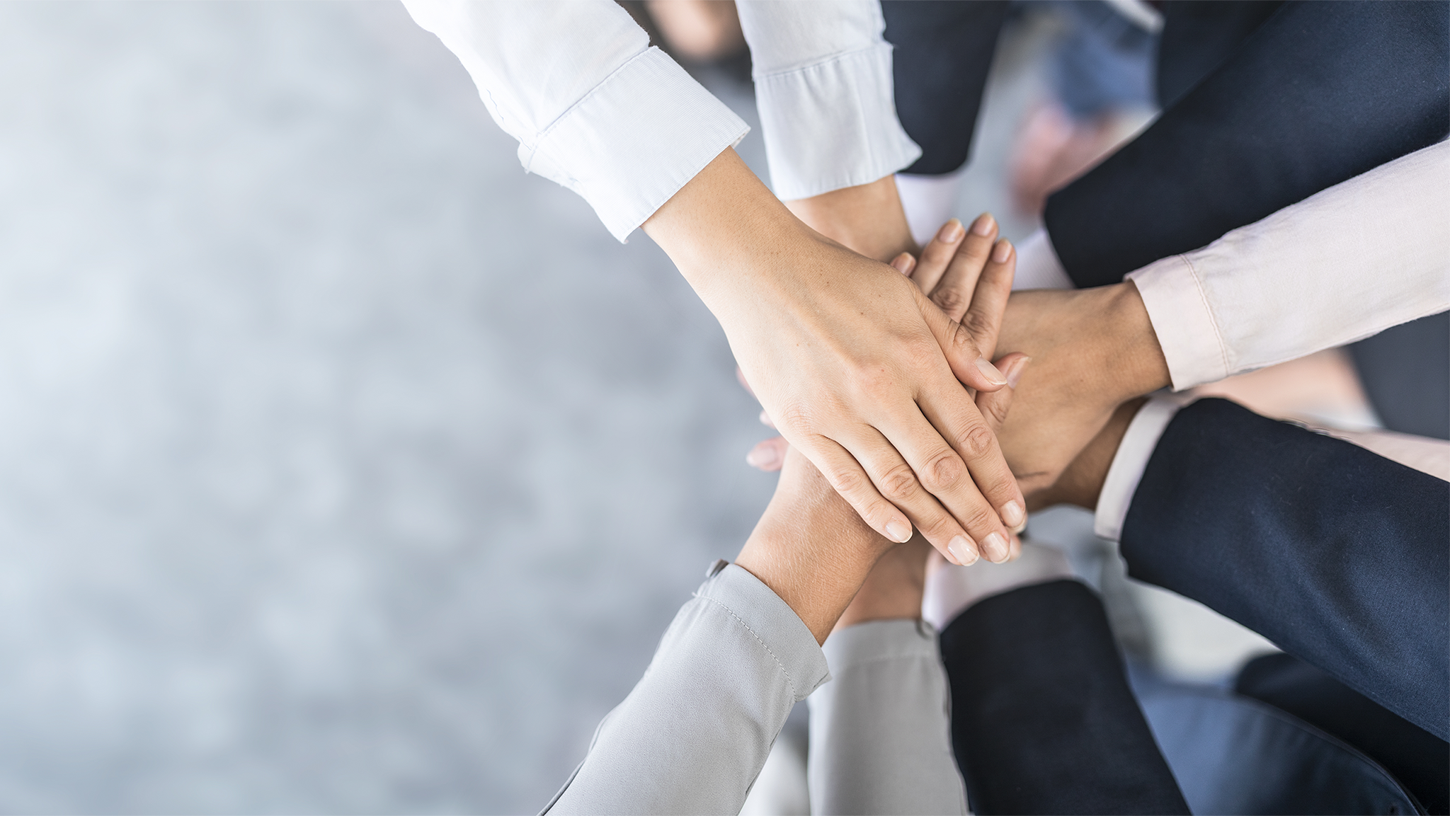 A group of diverse people place their hands together in a stack, symbolizing teamwork and unity. The background is a soft, blurred gray, emphasizing the focus on the hands.