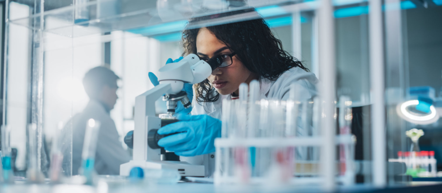 A scientist wearing a white lab coat and blue gloves examines a sample under a microscope in a laboratory. Test tubes are visible in the foreground, and another person works in the background.