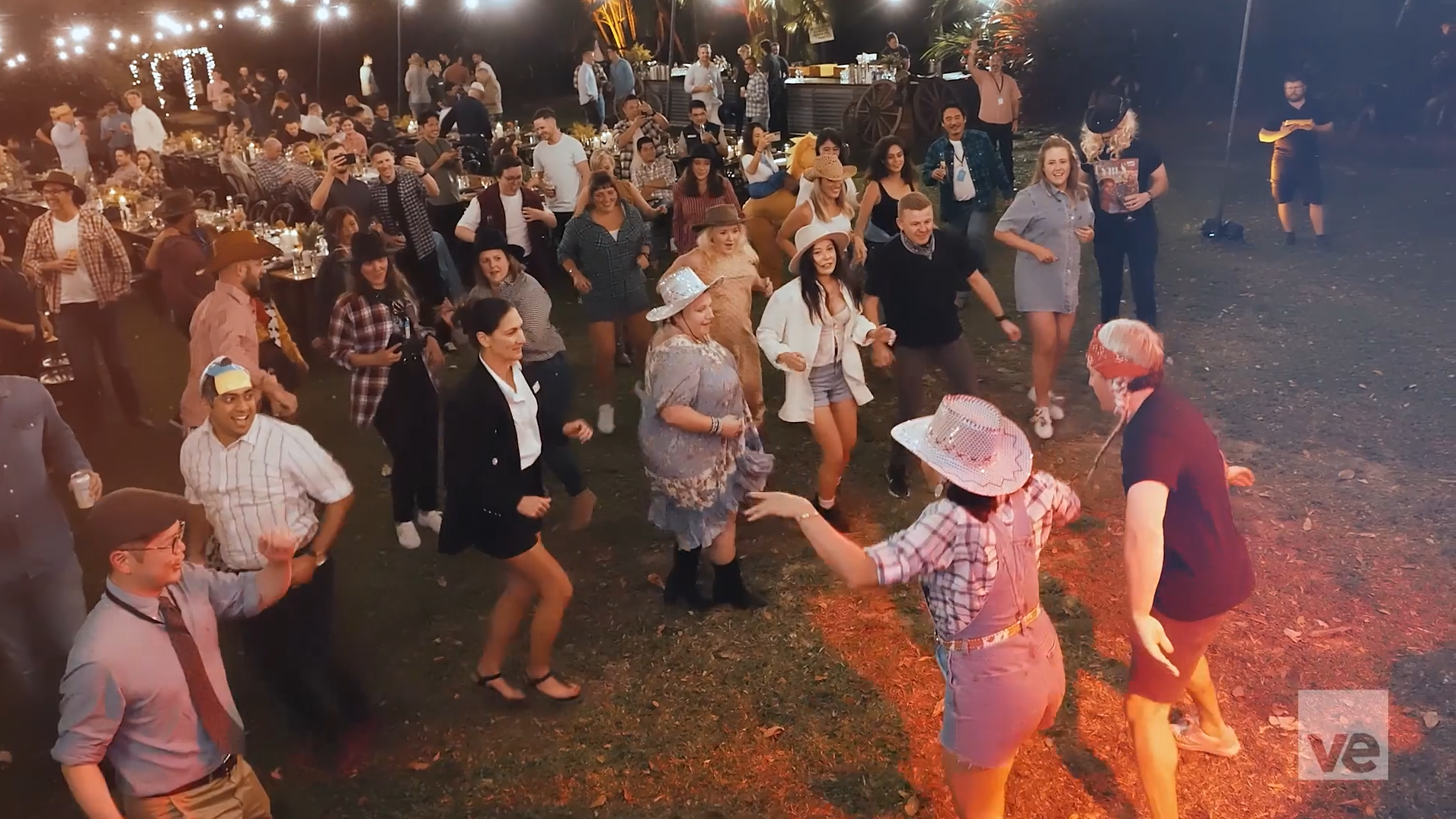 A lively outdoor gathering with people in casual and cowboy-themed attire dancing under string lights. A mix of cowboy hats and plaid shirts are visible, with participants enjoying the vibrant atmosphere on a grassy area.
