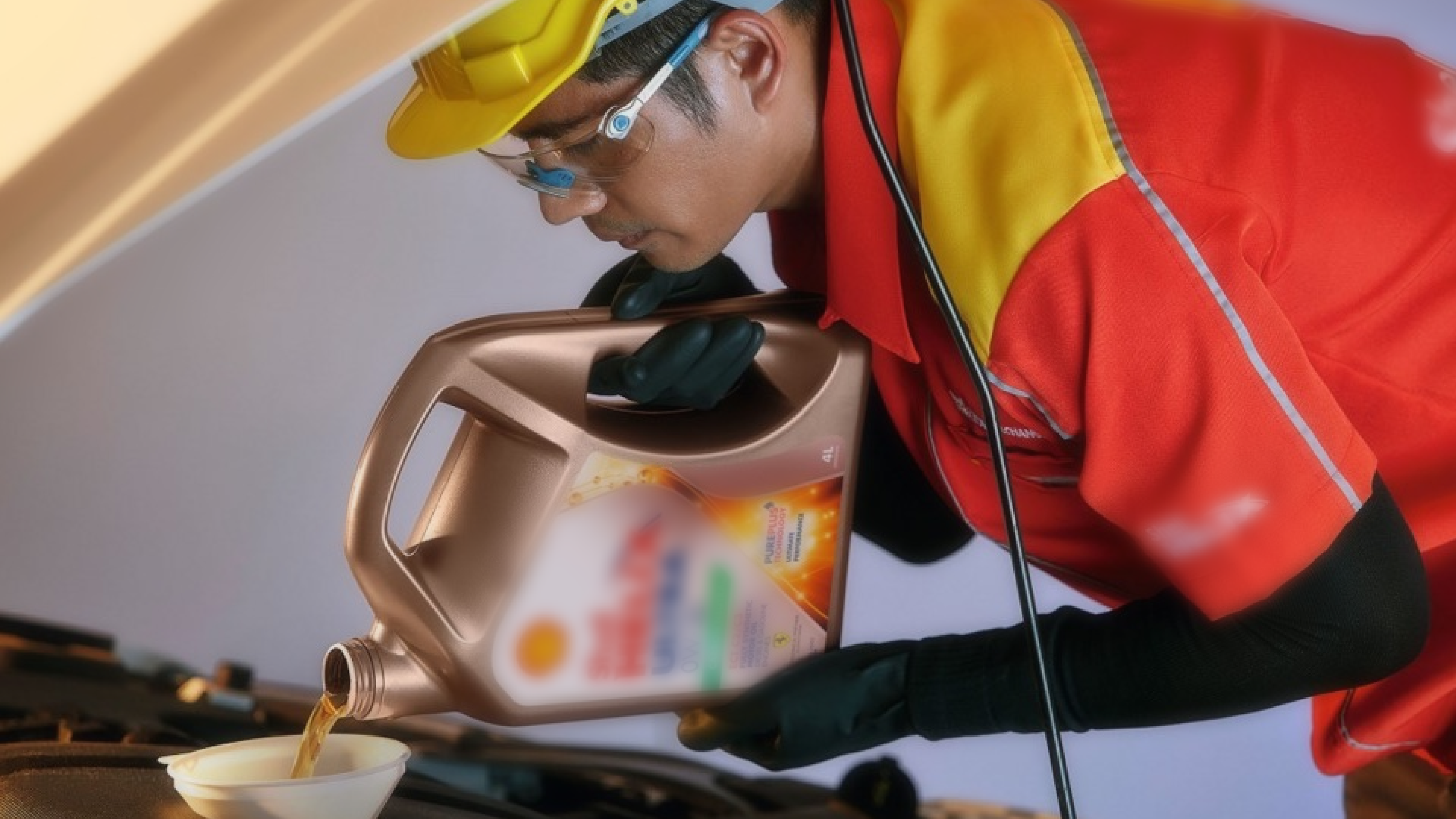 A worker in a red and yellow uniform, wearing protective eyewear and gloves, pours engine oil from a large bottle into a funnel under the hood of a vehicle.