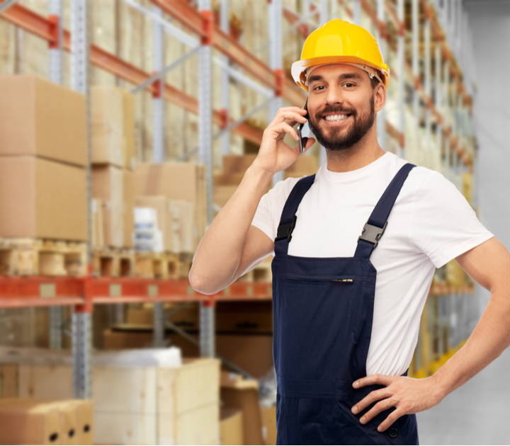 A man in a white t-shirt and blue overalls, wearing a yellow hard hat, smiles while talking on a smartphone. He stands in a warehouse with shelves stocked with boxes and packages in the background.