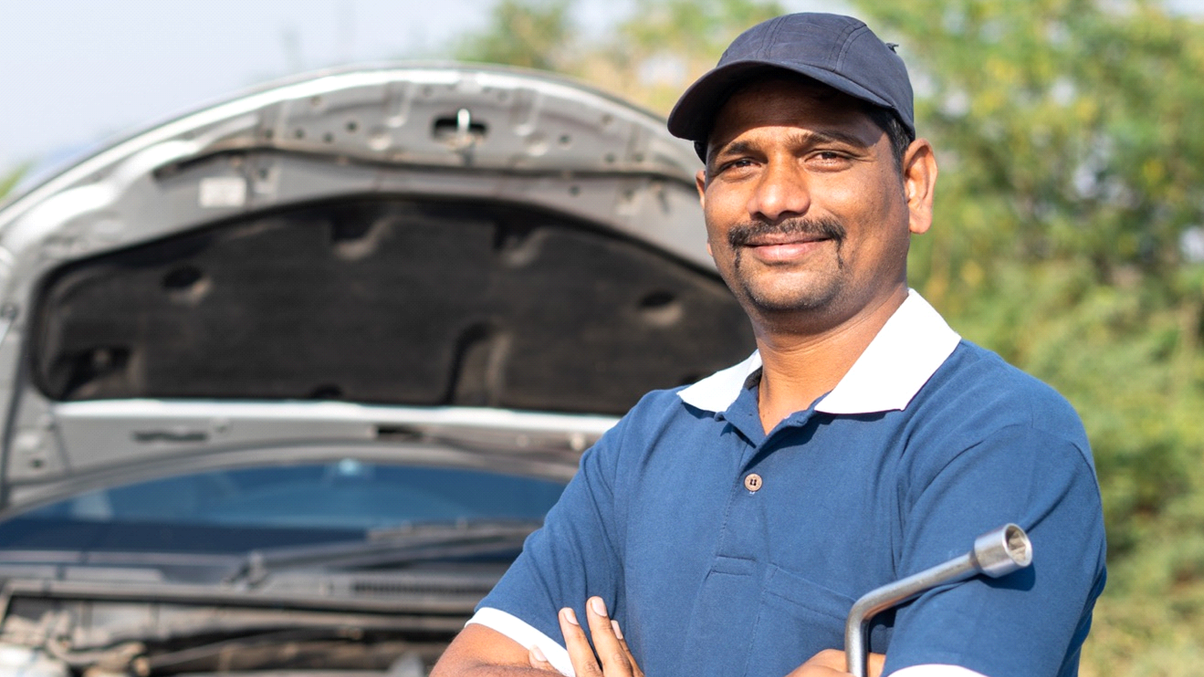 A man wearing a blue polo shirt and cap stands confidently with his arms crossed in front of an open car hood. Trees are visible in the background.