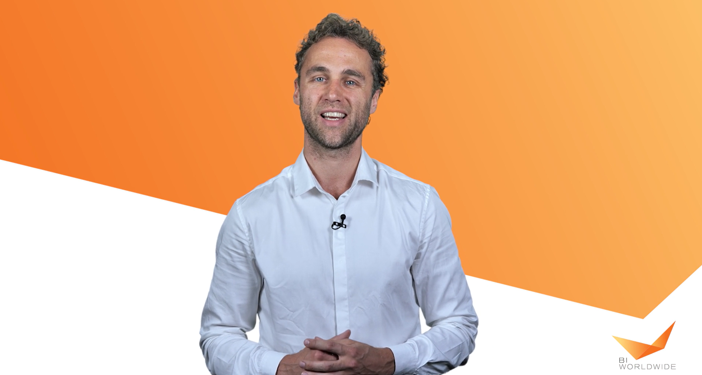 A man with curly hair smiles while clasping his hands. He wears a white button-up shirt. The background features an orange and white geometric design with a small BI WORLDWIDE logo in the bottom right corner.